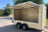 A cream-colored utility trailer with an open side cargo door and a raised metal canopy roof, parked outdoors on a paved surface with trees and blue sky in the background.
