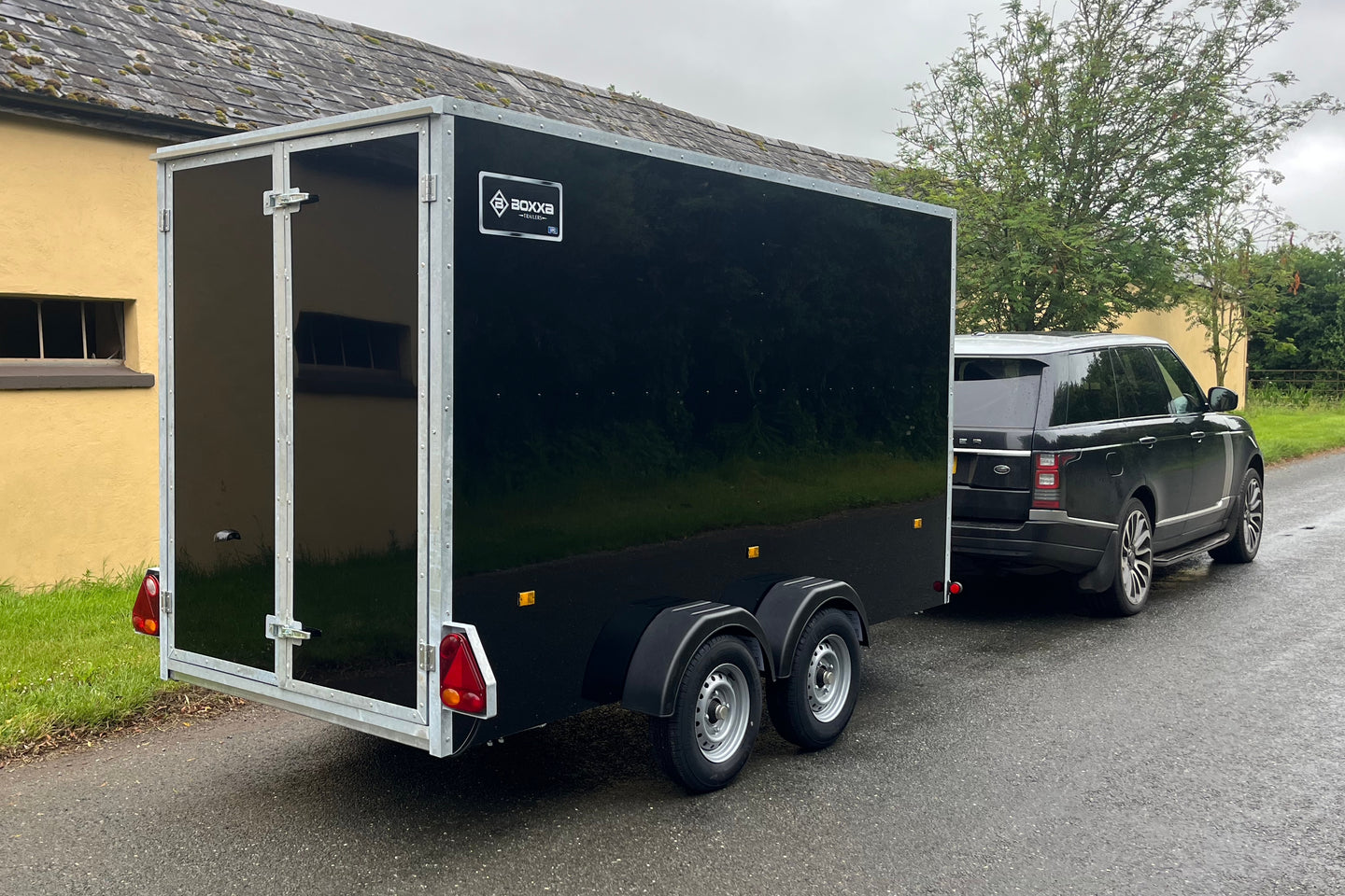 Black Boxxa trailer being towed by a car on a road with a building and trees in the background.