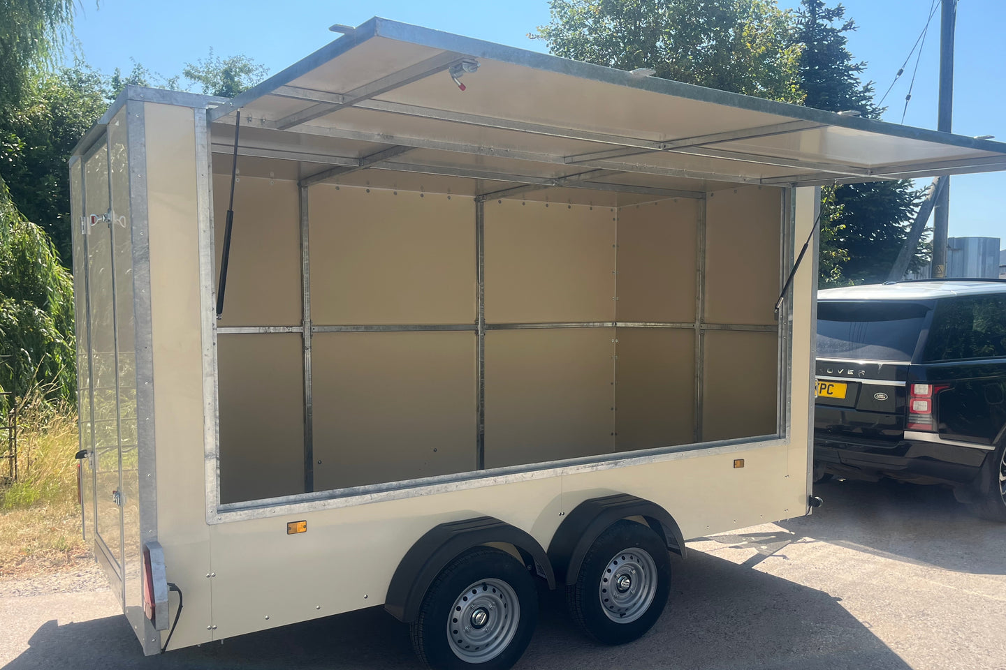 An empty cream-colored aluminum trailer with an open side door and extended roof canopy, parked outdoors next to a black SUV on a paved surface.