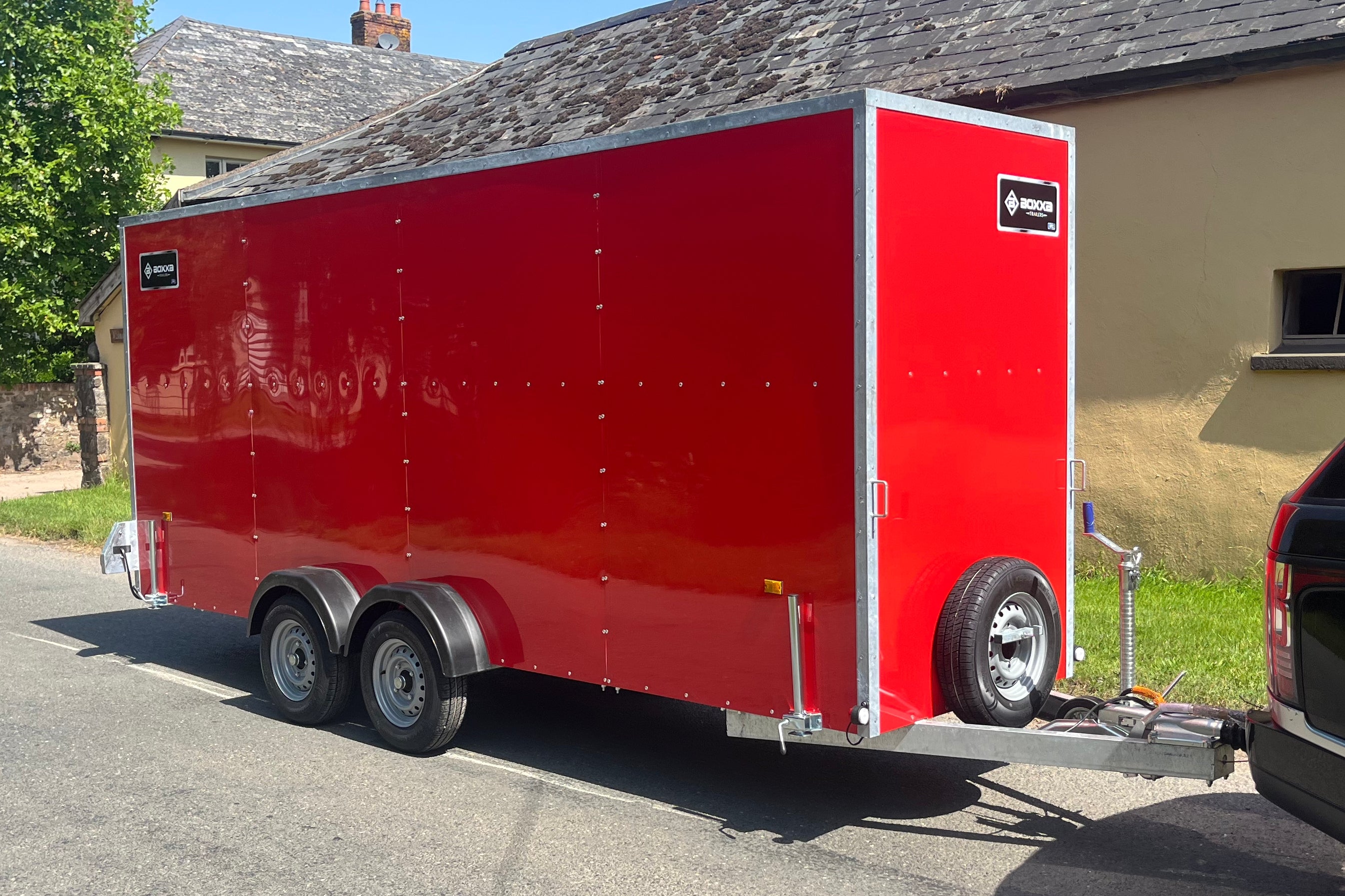 A red enclosed cargo trailer parked outdoors on a paved street, attached to a vehicle on the right side. The trailer has silver metal trim, dual rear wheels, a hitch, and a small black logo plate on the upper right side.