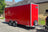 A red enclosed cargo trailer parked outdoors on a paved street, attached to a vehicle on the right side. The trailer has silver metal trim, dual rear wheels, a hitch, and a small black logo plate on the upper right side.