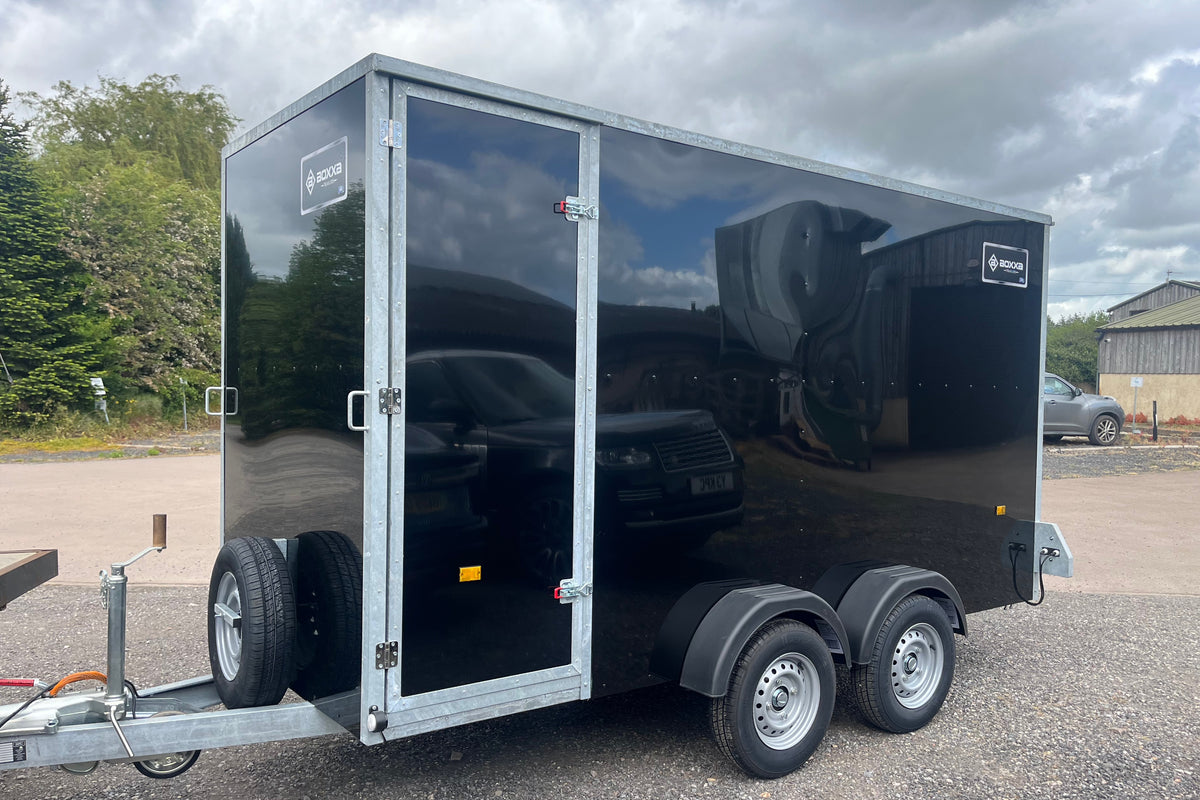 A black boxxatrailer with silver metal framing  he trailer has a side door and black exterior panels, with visible logos on the upper corners.
