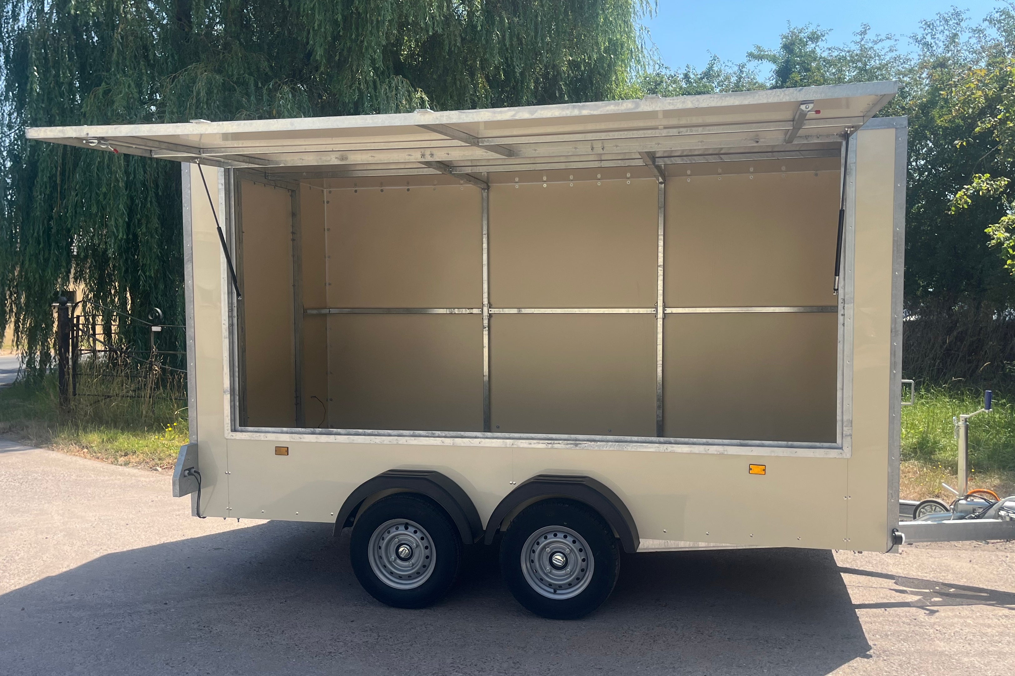 An empty beige aluminum trailer with an open side awning and interior, parked outdoors on a paved surface with trees in the background.
