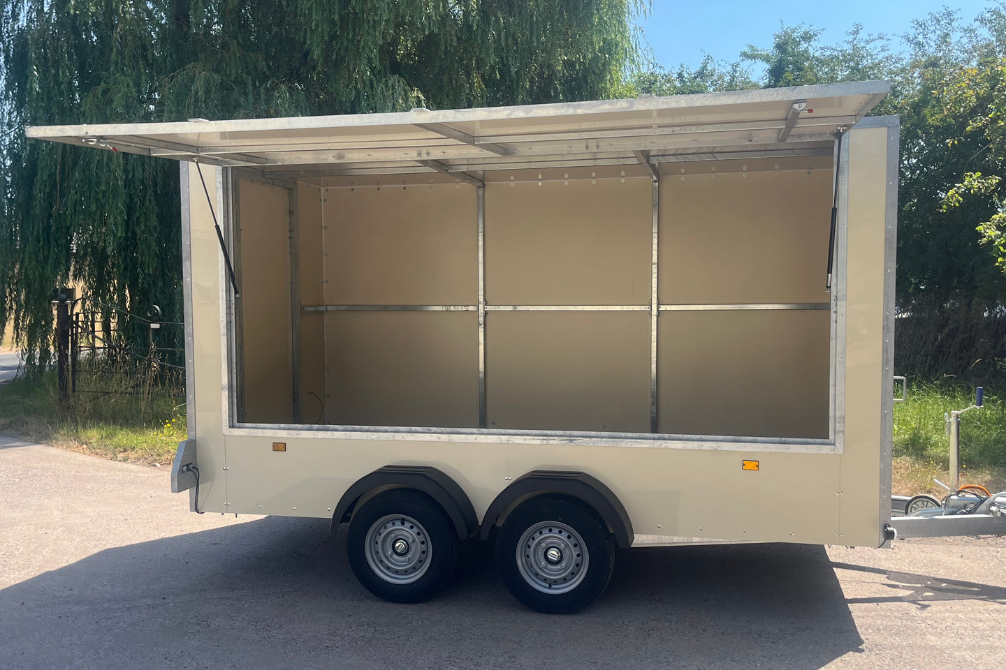 An empty beige aluminum trailer with an open side awning and interior, parked outdoors on a paved surface with trees in the background.