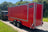 A red enclosed cargo trailer with silver trim and dual rear wheels, parked outdoors on a paved road next to grass and trees. A black vehicle is partially visible on the left, and a black logo plate reading 'BOXXA' is mounted on the side of the trailer.
