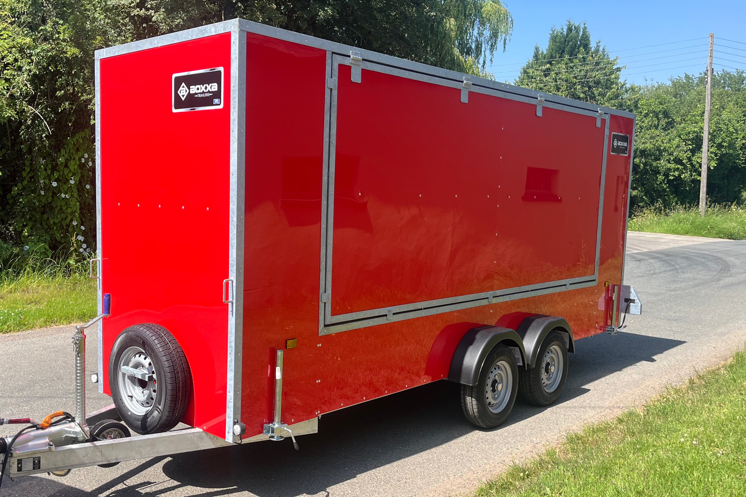 A red enclosed cargo trailer parked outdoors on a paved road with grass and trees on both sides. The trailer has silver trim, dual rear wheels, a hitch at the front, and a visible BOXXA logo on the upper left side.