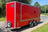 A red enclosed cargo trailer parked outdoors on a paved road with grass and trees on both sides. The trailer has silver trim, dual rear wheels, a hitch at the front, and a visible BOXXA logo on the upper left side.
