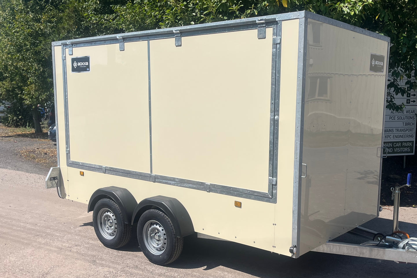 A cream-colored enclosed trailer with silver metal trim and black tires, parked outdoors on a paved surface with trees in the background.