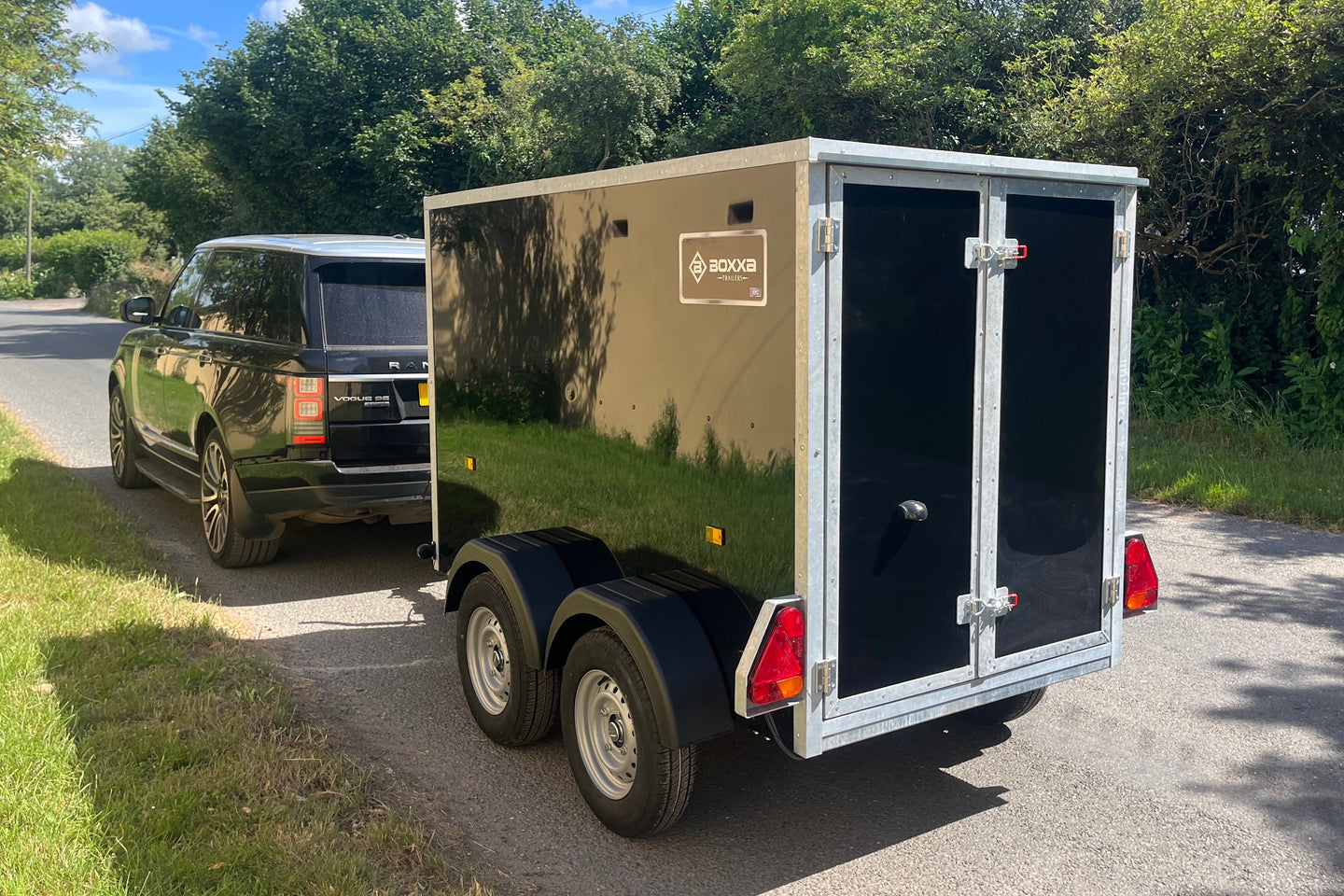 rear view of a black boxxa with twin barn doors being towed by a black vehicle on a country road.