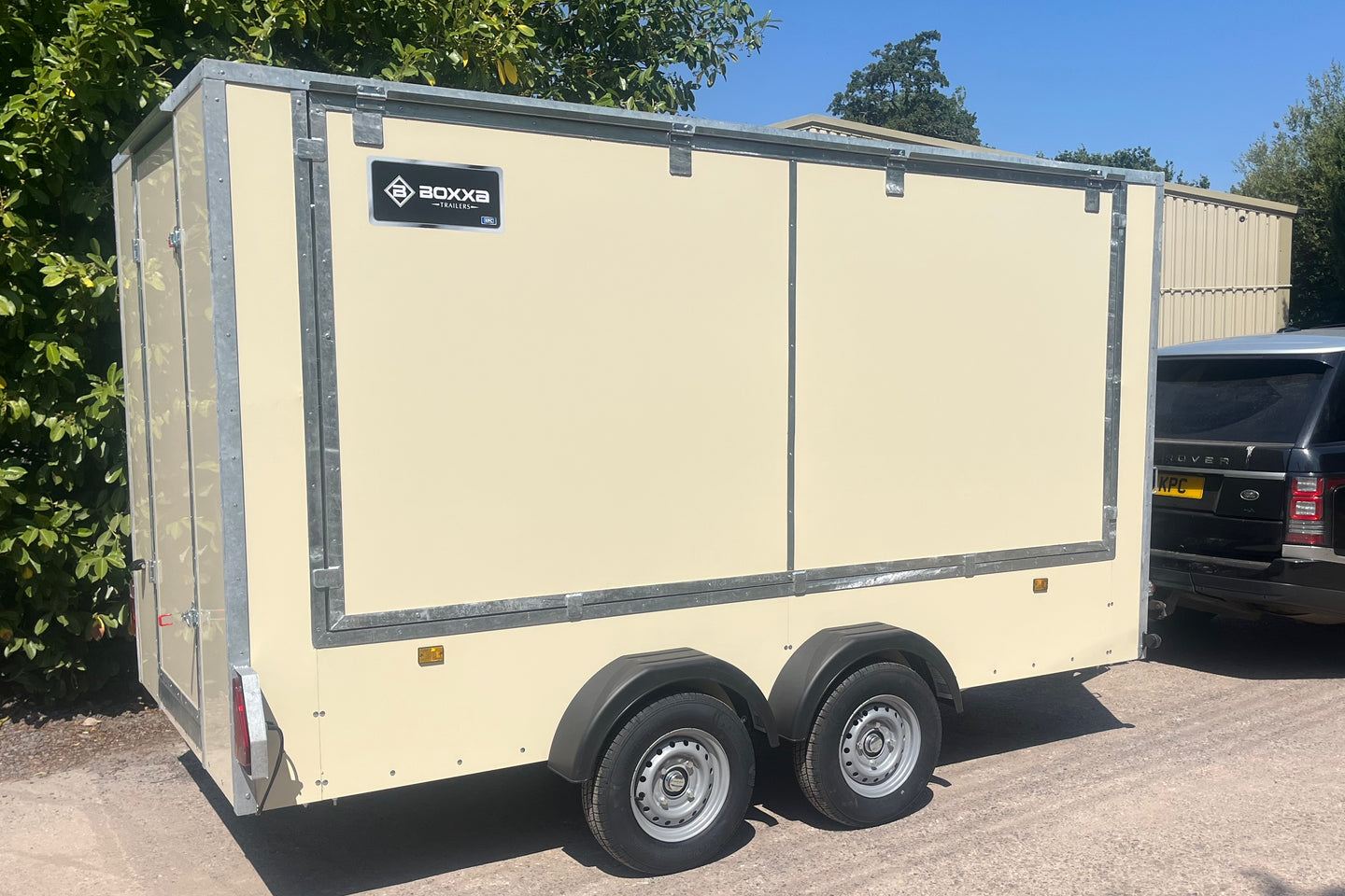 A cream-colored enclosed cargo trailer with silver trim and dual rear wheels, parked outdoors on a paved surface next to a black vehicle. The trailer features a black logo plate on the upper left side.