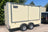 A cream-colored enclosed cargo trailer with silver trim and dual rear wheels, parked outdoors on a paved surface next to a black vehicle. The trailer features a black logo plate on the upper left side.