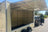 An empty beige cargo trailer with its side cargo door open, parked outdoors next to a black SUV on a paved road.