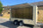 A beige enclosed utility trailer with its side cargo door open, parked outdoors on a paved surface in front of a tan building under a blue sky.