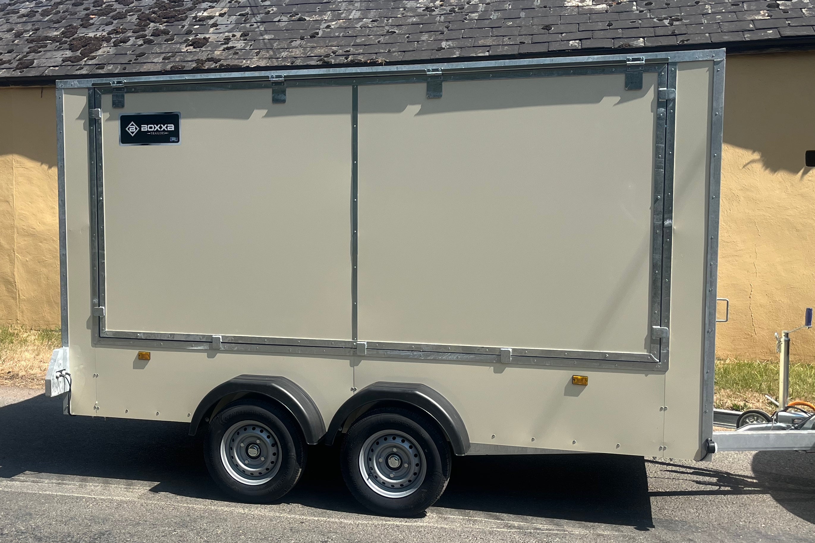 A beige enclosed cargo trailer with silver metal trim and dual wheels, parked outdoors against a tan building wall.