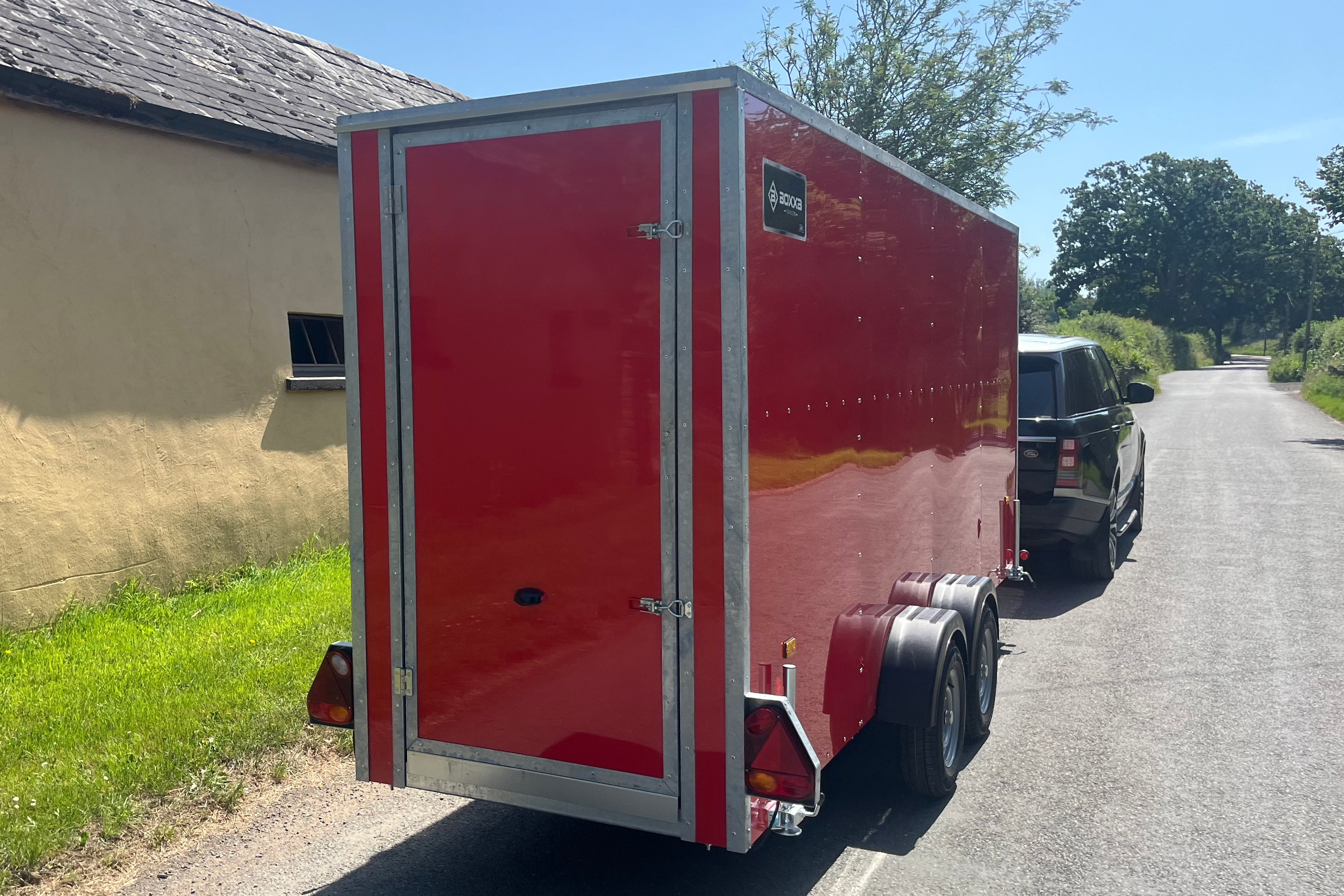 A red enclosed cargo trailer parked outdoors on a paved road, attached to a black vehicle. The trailer has silver metal trim and a rear double-door design with visible wheels and reflectors.
