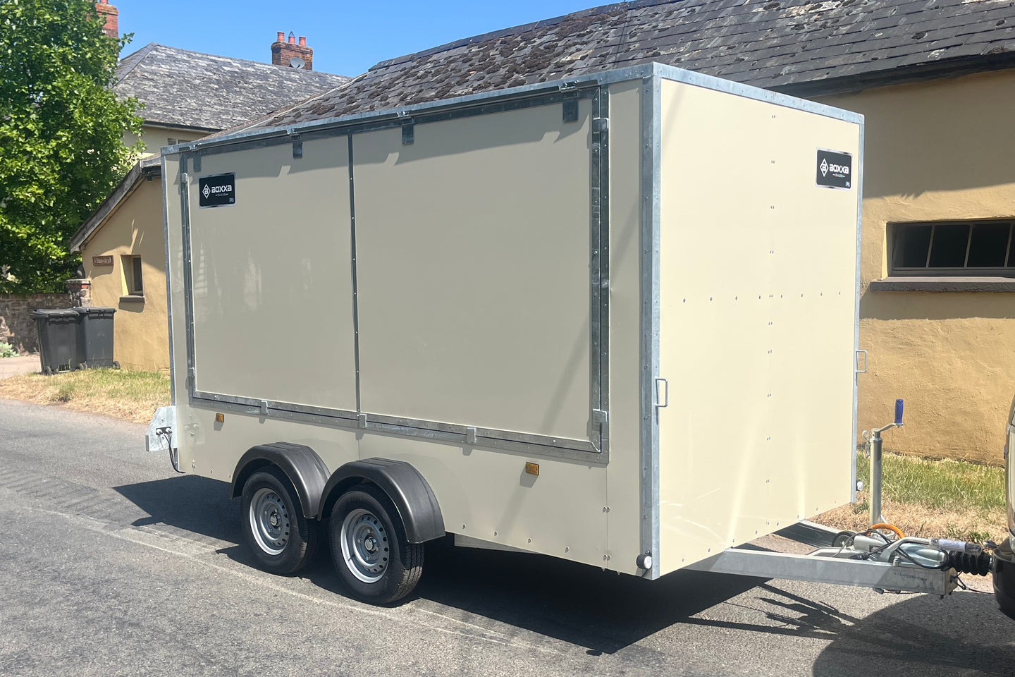 A beige enclosed trailer parked outdoors on a paved street in front of a house. The trailer has silver metal trim, dual rear wheels, and a hitch at the front right.
