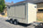 A beige enclosed trailer parked outdoors on a paved street in front of a house. The trailer has silver metal trim, dual rear wheels, and a hitch at the front right.