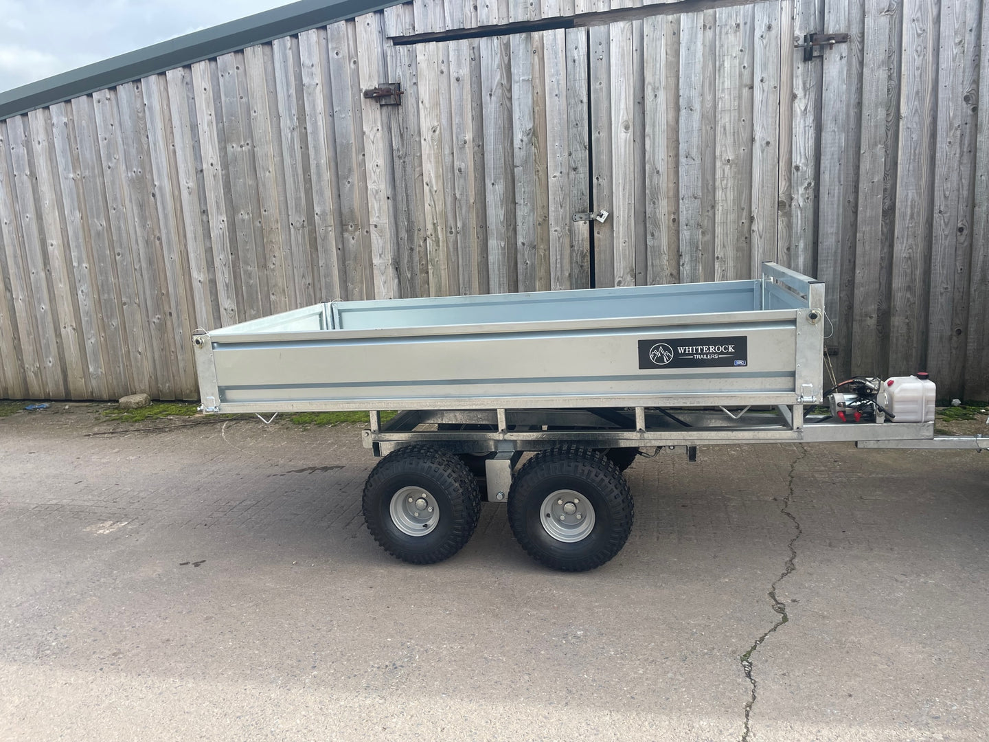 Side view of a galvanised steel twin axle trailer with low sides and tipping mechanism mounted at the front of the chassis.