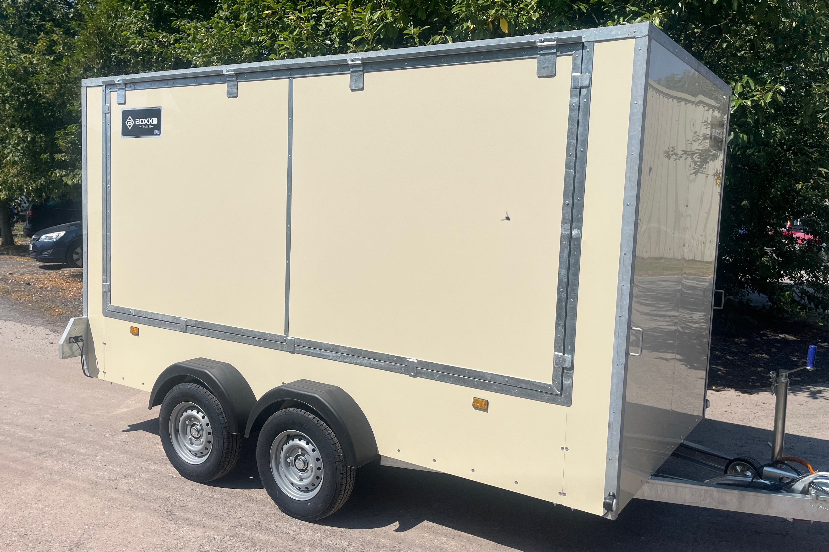 A cream-colored enclosed cargo trailer with silver metal trim and wheels, parked outdoors on a paved surface with trees in the background.